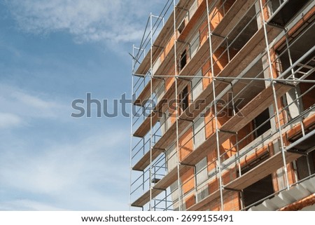 Construction site facade framed by metal scaffolding and exposed orange brickwork, concrete floors and clear blue sky background.