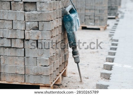 A construction site features stacks of gray bricks arranged neatly on wooden pallets. A drill is positioned against a pile of bricks, ready for use in the building process.