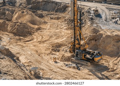 Construction site featuring drilling rig, heavy machinery, earthmoving activities, sand terrain, vertical drilling arm, tire tracks, construction equipment, wet ground, sunny day lighting
