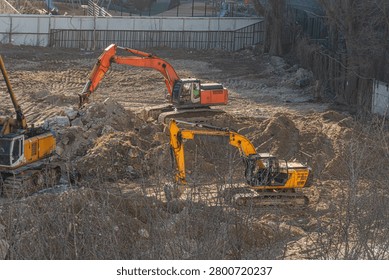 Construction site featuring excavators moving earth, orange and yellow machinery, industrial activity contrasting serene park environment beyond metal fence, bare trees indicating autumn or winter