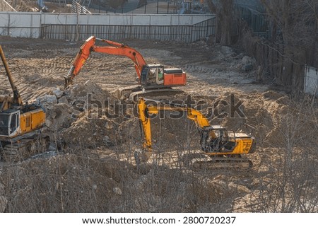 Construction site featuring excavators moving earth, orange and yellow machinery, industrial activity contrasting serene park environment beyond metal fence, bare trees indicating autumn or winter