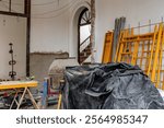 A construction site with a large black tarp covering a cement mixer. The scene is messy and disorganized, with tools and equipment scattered around. Scene is chaotic and potentially dangerous