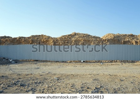 A construction site in Riyadh shows a gray metal fence with a large mound of dirt behind it. The ground is bare and there are some scattered items nearby. The sky is clear and bright.