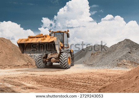Construction site shows a wheel loader transporting sand in a bucket. The loader operates among piles of earthy materials against a cloudy sky.