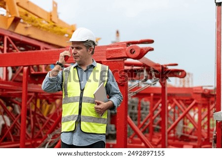 Construction site supervisor or project manager wearing a white hard hat and safety vest using a walkie-talkie while holding a tablet, standing near red tower crane equipment.