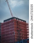 Construction site with a tall building covered in red scaffolding and a crane against a cloudy sky in Leeds, UK.