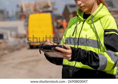 Construction site worker monitors project progress using a tablet in bright safety gear during the day