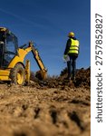 A construction supervisor in a hard hat and reflective vest stands on a muddy site holding documents while watching an excavator during earthmoving work.