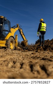 A construction supervisor in a hard hat and reflective vest stands on a muddy site holding documents while watching an excavator during earthmoving work.