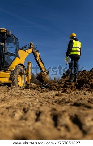 A construction supervisor in a hard hat and reflective vest stands on a muddy site holding documents while watching an excavator during earthmoving work.