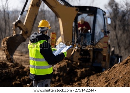 A construction supervisor holds documents and communicates with an excavator operator during active trench excavation on a worksite.