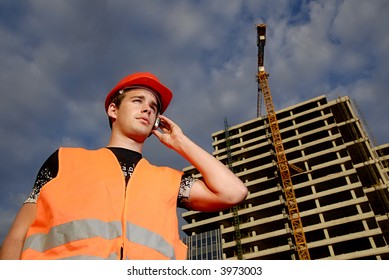 Construction Supervisor In Safety Helmet And Reflex Vest Talking On The Phone In Front Of Construction Site.