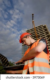 Construction Supervisor In Safety Helmet And Reflex Vest With Notebook In Front Of Construction Site.