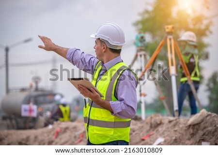 Construction survey engineer wear safety uniform under inspection and survey workplace by tablet with excavation truck digging, theodolite and worker construction road background.