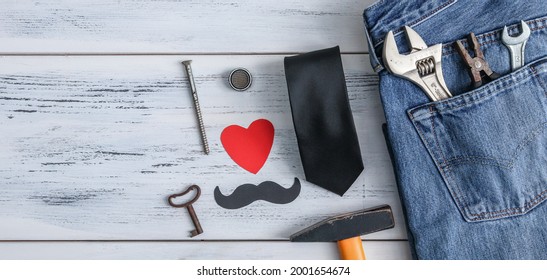 Construction Tools, Jeans, A Tie And A Paper Heart With A Mustache Lie On The Right On A White Wooden Table With Space For Text On The Left, Close-up Top View.