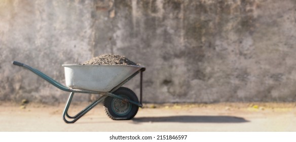 construction wheelbarrow with a pile of sand. a dirty construction wheelbarrow stands against a gray wall in the sun