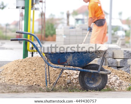 Construction wheelbarrow standing beside gravel and concrete curb stones at building site. Worker in background