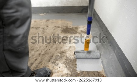 Construction worker applying a gray waterproofing membrane with a paint roller on a concrete floor, ensuring protection against moisture and leaks