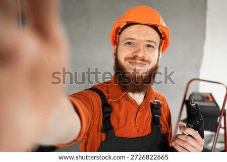 Construction worker bearded man in hardhat making selfie with drill at construction site. Repairman making house repair. Professional builder indoors. copy space