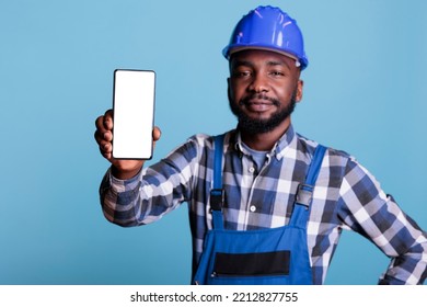 Construction worker in blue uniform work coveralls holds cell phone with empty screen for advertising isolated on blue background. Male professional builder in hard hat looking at camera. - Powered by Shutterstock