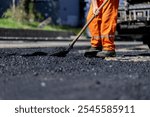 A construction worker in bright orange attire is evenly distributing asphalt on a road site, under clear skies and bright sunlight, ensuring smooth pavement.