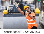 A construction worker in a bright safety vest and hard hat is signaling for vehicles to stop while overseeing roadwork in a busy urban area.