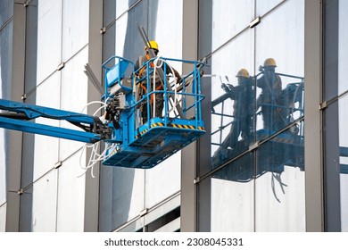 A construction worker in the bucket of a crane is installing insulation in the window block of the building. A man in the cradle, dismantling the old insulation of a sealed gasket to a new one. Worker