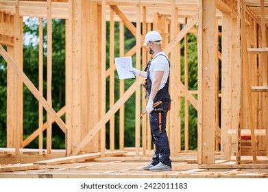 Construction worker building two-story wooden frame house. Man with beard, wearing jumpsuit and hard hat, reviewing the building plan. Intention to implement modern ecological construction practices. - Powered by Shutterstock