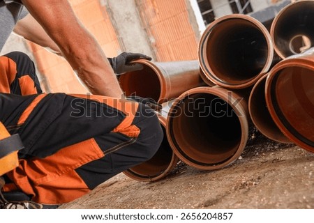 A construction worker is carefully arranging plastic pipes on the ground at a building site. The setting is busy with ongoing construction activities around.