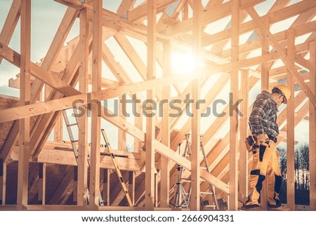 A construction worker is carefully assembling a wooden frame under the bright afternoon sun. Ladders and tools are scattered around the active site.