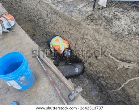 Construction worker carefully cuts large embedded rock in narrow trench while avoiding exposed tree roots during sensitive underground utility excavation.

