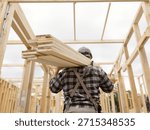 A construction worker carries a stack of lumber on his shoulders inside the wooden frame of a new house under construction.