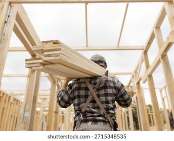 A construction worker carries a stack of lumber on his shoulders inside the wooden frame of a new house under construction.
