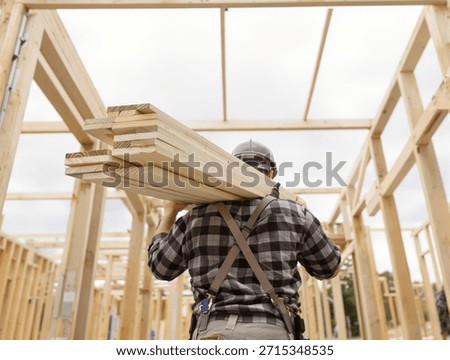 A construction worker carries a stack of lumber on his shoulders inside the wooden frame of a new house under construction.