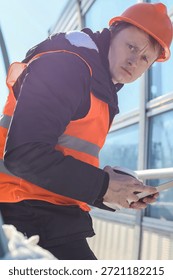 Construction worker checking digital tablet on building site. Worker in safety gear examines tablet device outdoors near modern glass building on a sunny day