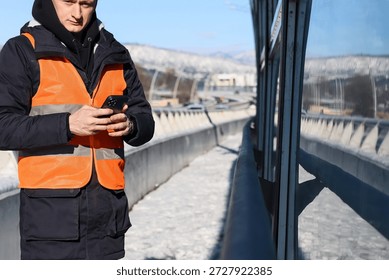 Construction worker checking phone on winter bridge. Man in orange safety vest and helmet using smartphone on snowy bridge with scenic mountain view