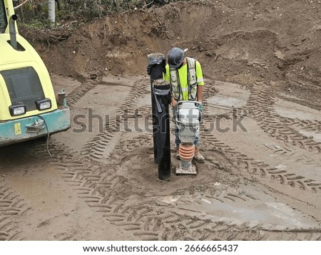 Construction worker compacts soil around underground valve with jumping jack compactor while roller compactor levels trench backfill on watermain installation site.