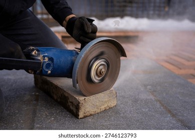 Construction worker cutting concrete curb with angle grinder, rotating abrasive disc trimming paving stone for sidewalk installation and urban landscaping work.