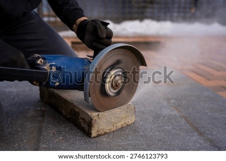 Construction worker cutting concrete curb with angle grinder, rotating abrasive disc trimming paving stone for sidewalk installation and urban landscaping work.