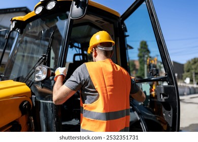 A construction worker dons safety gear and prepares to enter a large piece of machinery at a bustling construction site under clear blue skies.