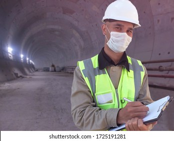 Construction Worker Dressed In Medical Mask, Uniform And White Hardhat Stands With Clipboard At Metro Tunnel Under Construction