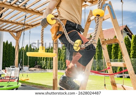 A construction worker is engaging in the installation of new playground equipment in an outdoor recreation area. Wearing gloves and sturdy boots, the worker is seen climbing a wooden ladder