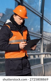 Construction worker examines clipboard while standing near reflective glass wall in winter.. Inspector in high-visibility clothing checks records on a clipboard in snowy outdoor environment.