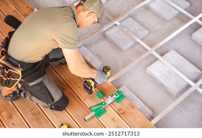 A construction worker is focused on installing patio flooring with power tools on a wooden deck. The setting is a building site filled with construction materials.