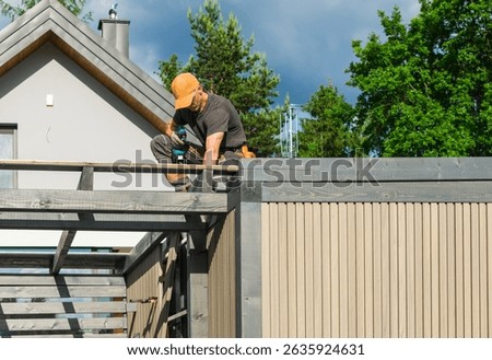 A construction worker is focused on installing elements on the roof of a residential building during a bright, sunny day. Trees and clouds provide a backdrop.