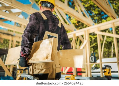A construction worker in a hard hat and gloves transports wooden planks at a sunny building site, showcasing the active phase of construction work.