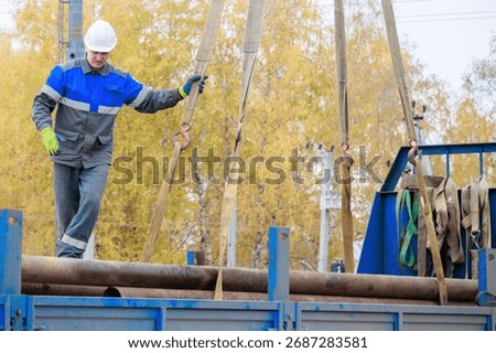 A construction worker in a hard hat and gloves maneuvers steel pipes on a truck. The setting is an outdoor construction site during autumn.