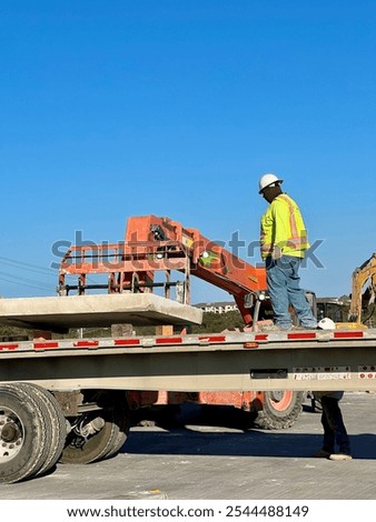 A construction worker in a hard hat and high-visibility vest stands on a flatbed trailer guiding a forklift as it lifts a large concrete slab. Blue sky and industrial equipment set the scene.