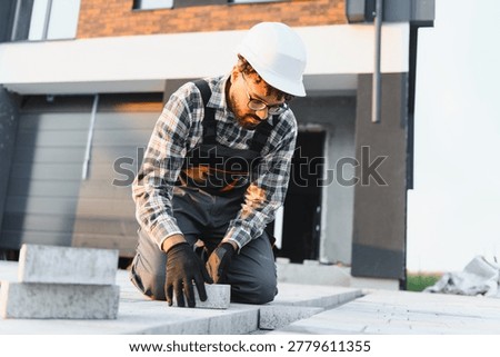 Construction worker in hard hat laying paving stones, creating an outdoor pathway for a new modern house