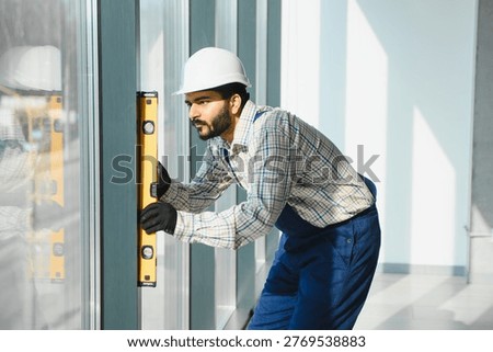 Construction worker in hard hat and overalls measuring window frame alignment with a spirit level, ensuring precision during installation